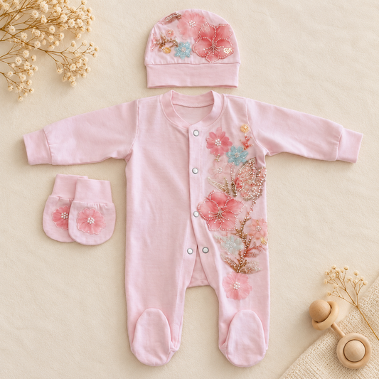 A baby footed onesie set in light pink, featuring embroidered floral patterns in pink, gold, and light blue. The set includes a long-sleeve onesie with snap closures, a matching bonnet, and a pair of booties, arranged on a beige fabric background with dried flowers and a wooden rattle.