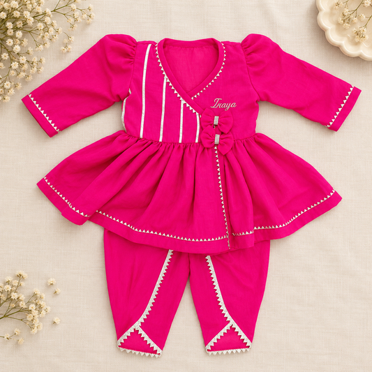 A bright pink baby outfit set consisting of a long-sleeve peplum-style top and matching pants, laid flat on a light beige fabric background with small white flowers as decorative props. The top has white vertical stripes, white trim, a bow detail, and the name 'Traya' embroidered on the chest.