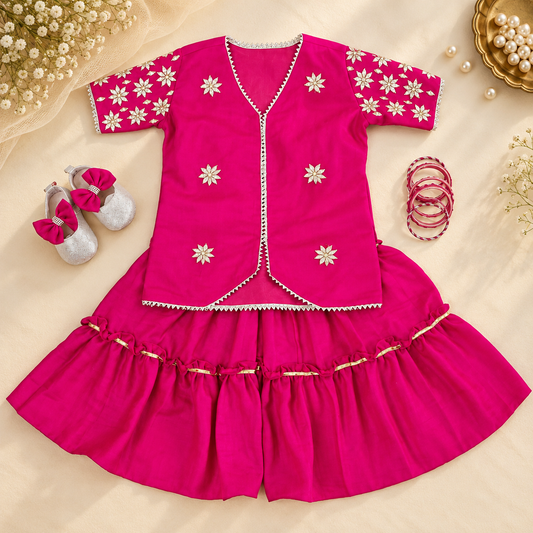 A bright pink traditional outfit for a baby girl, featuring a short-sleeve embroidered vest and matching ruffled pants, styled in a flat lay with silver baby shoes, red bangles, and decorative flowers on a beige fabric background.