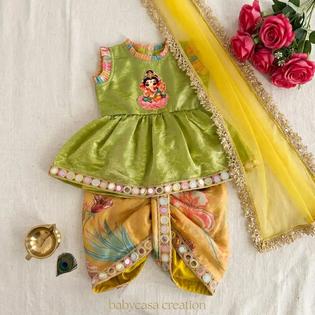 A traditional Indian baby outfit set featuring a lime green sleeveless top with a Ganesha embroidery, mustard yellow patterned pants, and a yellow dupatta with gold trim, styled on a white textured background with decorative props.