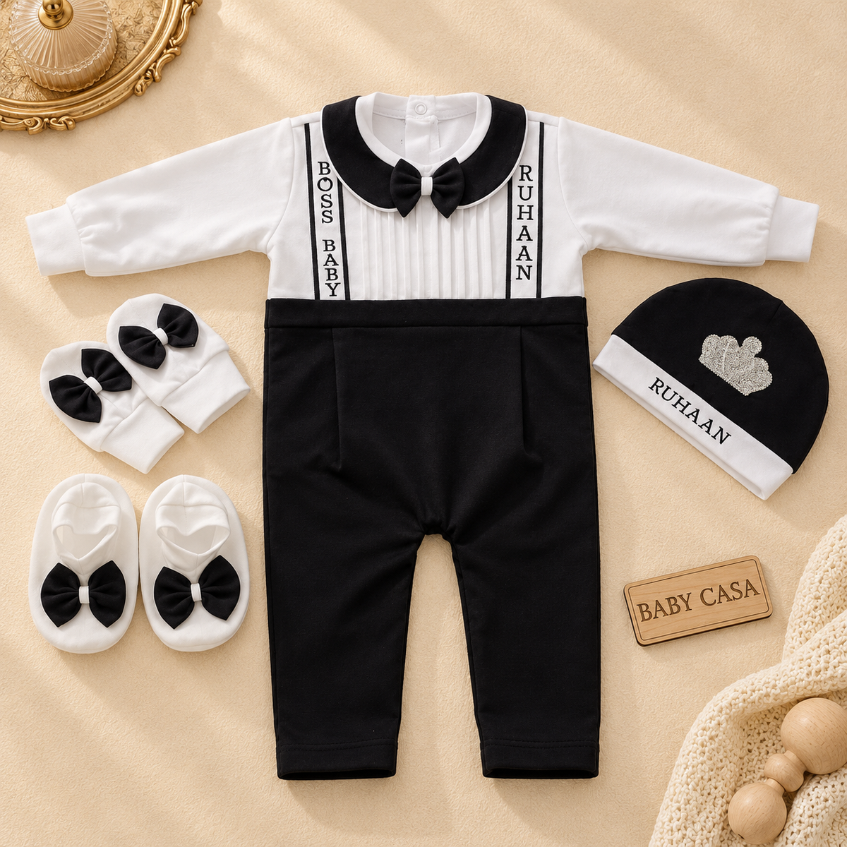 A baby outfit set laid flat on a beige fabric background, including a long-sleeve black-and-white romper with a black bow tie and suspenders, black pants, white booties with black bows, a black-and-white beanie, and a small wooden sign reading BABY CASA.