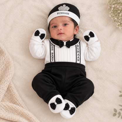 A baby lying on a soft cream blanket wearing a black and white formal outfit with a matching hat, mittens, and booties. The outfit includes a white long-sleeve top with black bow tie and suspenders, black pants, and a black hat with a white band reading RUHAAN.