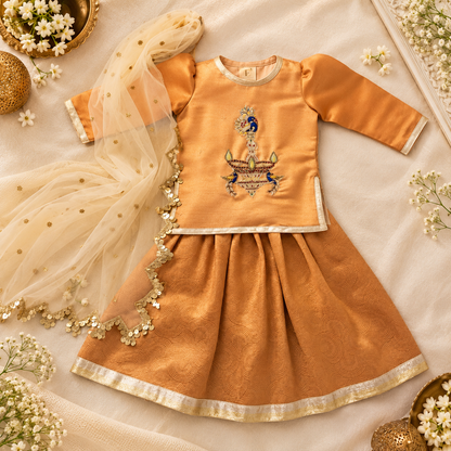 A traditional Indian children's outfit laid flat on a cream fabric background with decorative flowers and gold ornaments. The outfit consists of a golden-yellow long-sleeve top with a central embroidered motif, a matching pleated skirt with a gold border, and a sheer cream dupatta with gold dots and floral trim.