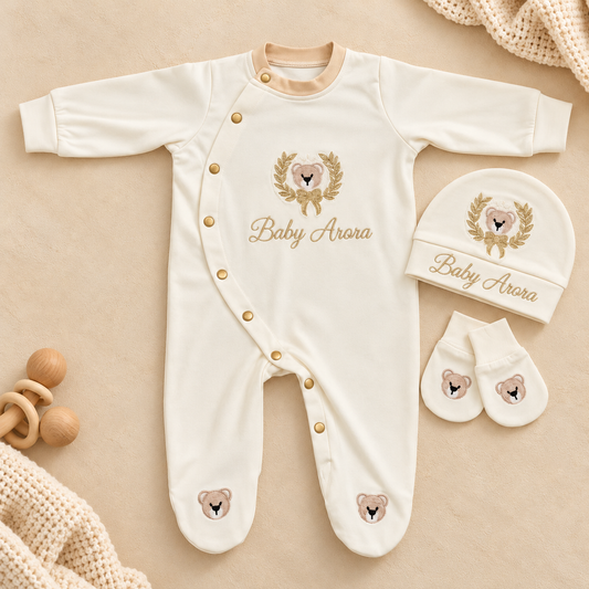 A baby footed onesie set in cream with gold buttons, featuring a teddy bear graphic and the text 'Baby Aurora' on the chest and hat. The set is styled in a flat lay with a matching beanie, mittens, a wooden teether, and soft knit blankets on a beige background.