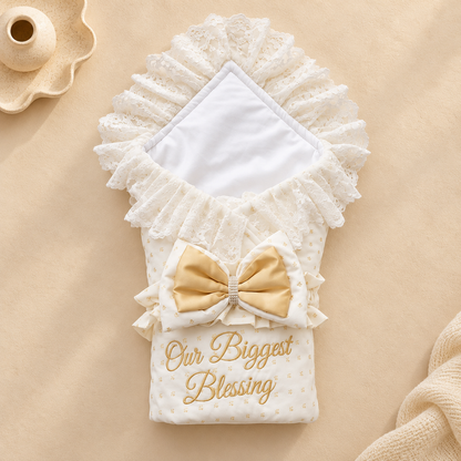 A folded baby blanket or swaddle with ruffled lace trim, a satin bow, and embroidered text reading 'Our Biggest Blessing' on a beige fabric background.