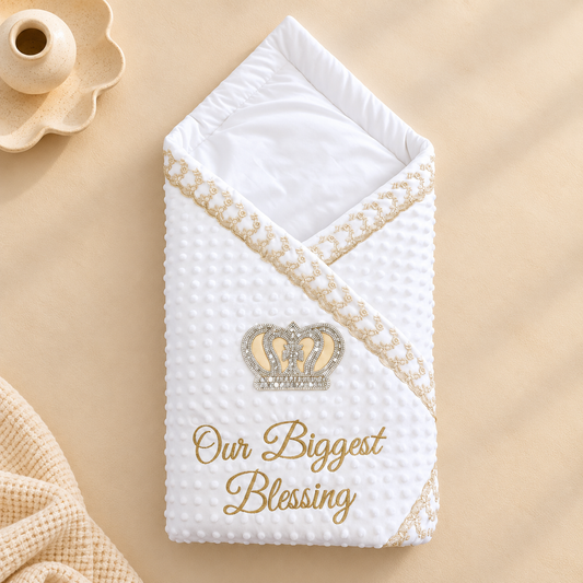 A white textured baby blanket or swaddle with a gold embroidered crown and the words 'Our Biggest Blessing,' styled on a beige surface with a small wooden candle holder and cream knit fabric nearby.
