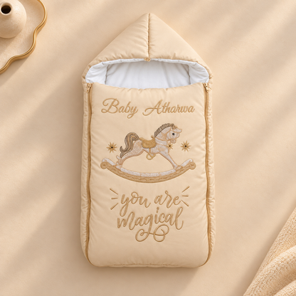 A beige quilted baby footmuff or sleeping bag with embroidered text reading 'Baby Atharwa' and 'you are magical' around a rocking horse illustration, laid flat on a matching beige fabric surface.