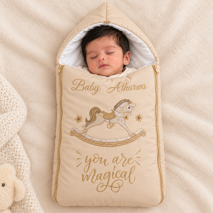 A sleeping newborn baby is swaddled in a beige hooded baby blanket bag with gold embroidery featuring a rocking horse and the text 'Baby Atharva' and 'you are magical'. The baby bag is placed on a soft beige blanket next to a knitted white blanket and a plush toy.