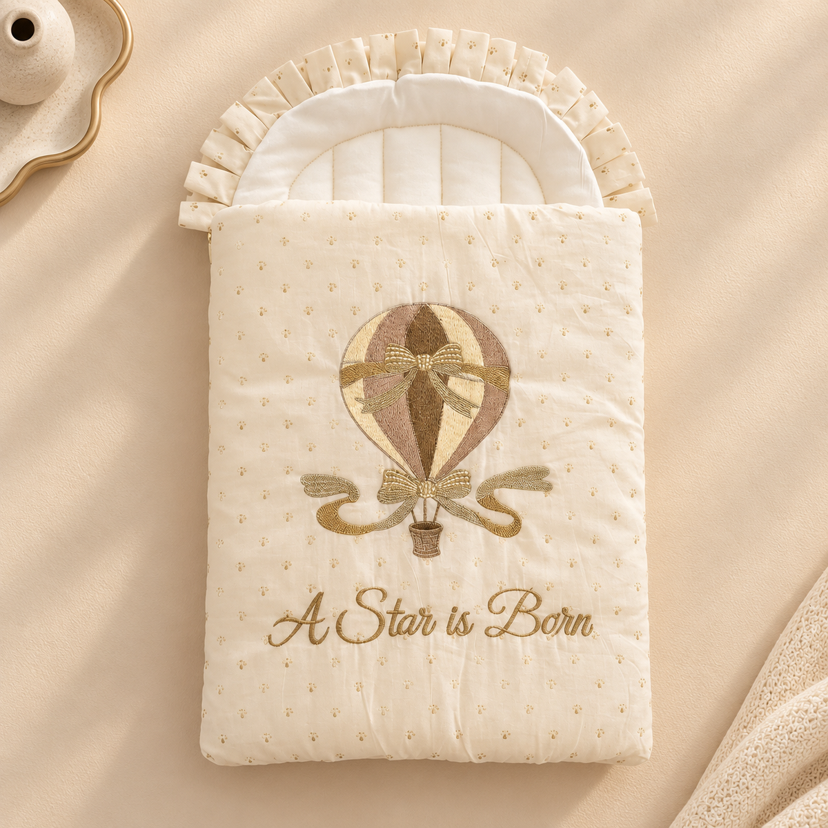 A cream-colored quilted baby blanket or swaddle with a hot air balloon embroidery and the text 'A Star is Born' in gold script. The blanket is styled flat-lay on a matching beige fabric background with a decorative tray partially visible in the top left corner.