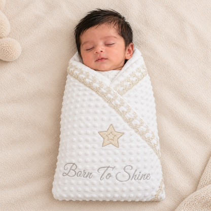 A sleeping newborn baby swaddled in a white textured minky dot blanket with a beige embroidered star and the words 'Born To Shine' on a soft beige blanket.