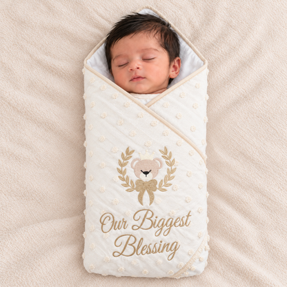 A sleeping newborn baby wrapped in a cream-colored quilted swaddle blanket with gold embroidered text reading 'Our Biggest Blessing' and a teddy bear design, lying on a soft beige blanket.