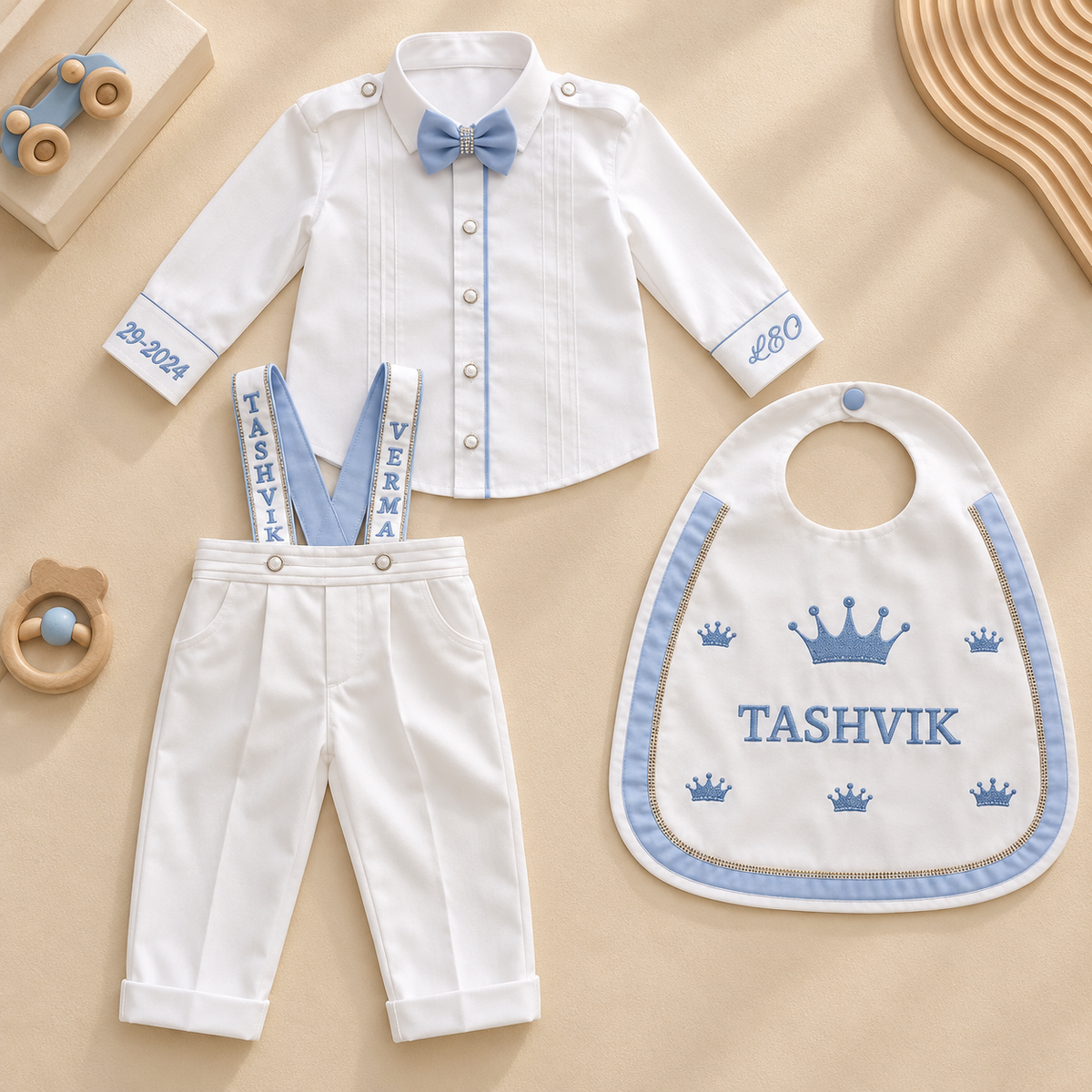 A baby boy's three-piece outfit set laid flat on a beige background, including a white long-sleeve button-up shirt with a light blue bow tie, white pants with light blue suspenders, and a white bib with blue crown and name embroidery.