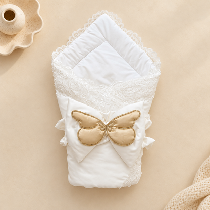 A folded white baby blanket or swaddle with lace trim and a large gold satin bow on the front, photographed on a warm beige background with small decorative props.