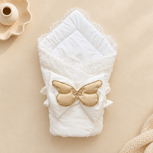 A folded white baby blanket or swaddle with lace trim and a large gold satin bow on the front, photographed on a warm beige background with small decorative props.