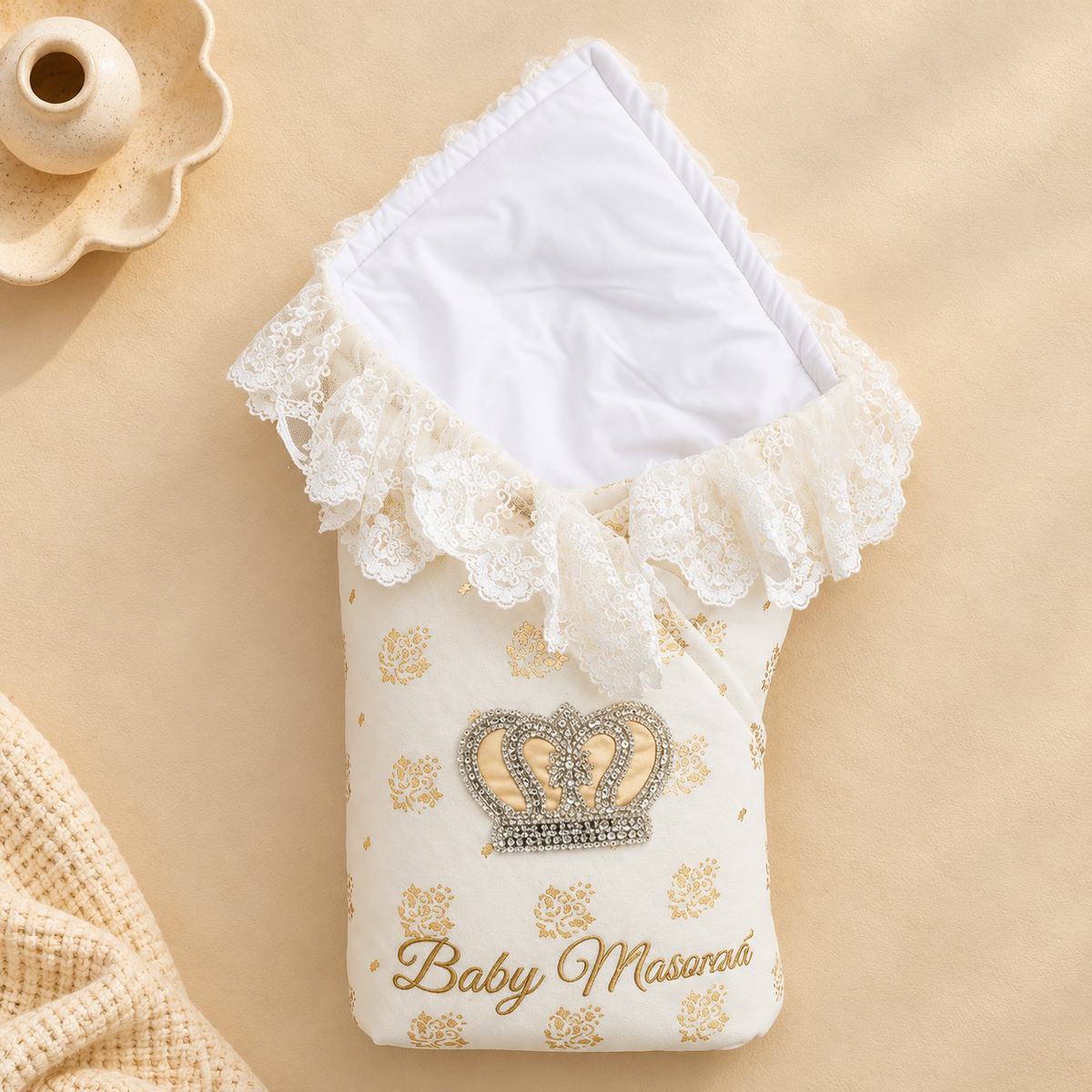 A baby swaddle blanket with a white lining, decorated with a lace-trimmed top, gold floral pattern, a jeweled crown motif, and the text 'Baby Masorah' on a beige background.