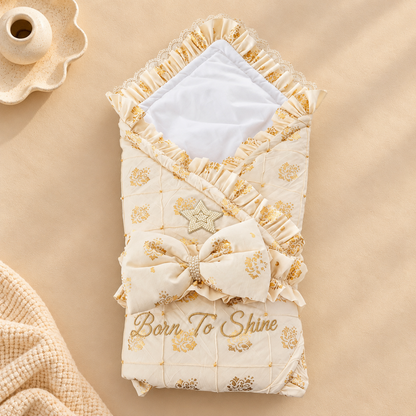 A cream and gold quilted baby swaddle blanket with ruffles, a matching bow, lace trim, and the words 'Born To Shine' embroidered on the front, styled on a beige background with a small decorative dish and textured fabric.