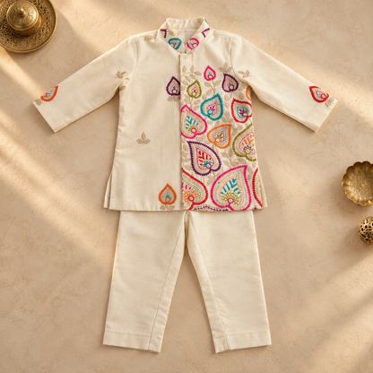 A cream-colored two-piece traditional outfit laid flat on a beige textured surface, featuring a long-sleeve jacket with colorful embroidered paisley patterns on the front and small motifs on the sleeves, paired with matching straight-leg pants. Decorative brass bowls are placed in the corners.