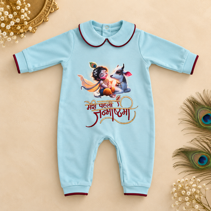 A light blue baby romper with a cartoon graphic of a child and a goat, featuring Hindi text and dark red trim, laid flat on a beige background with decorative peacock feathers and flowers.