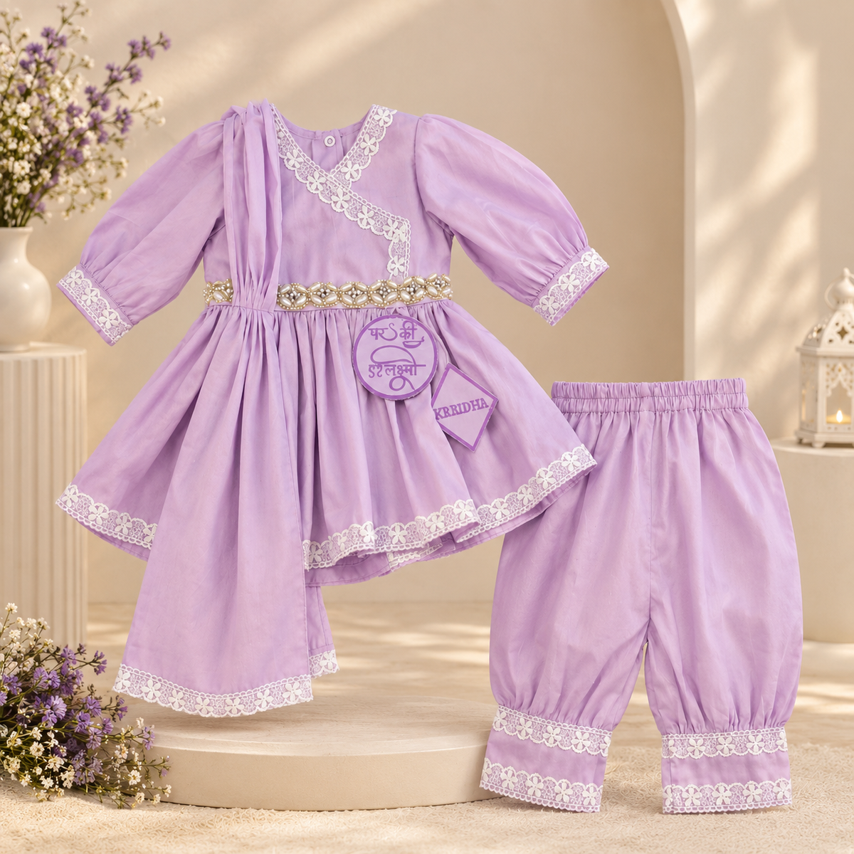 A baby clothing set in light purple, featuring a long-sleeve tunic top with white lace trim and a decorative gold-tone belt, paired with matching elastic-waist pants with lace cuffs, displayed on a beige set with floral props.