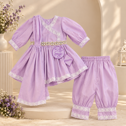 A baby clothing set in light purple, featuring a long-sleeve tunic top with white lace trim and a decorative gold-tone belt, paired with matching elastic-waist pants with lace cuffs, displayed on a beige set with floral props.