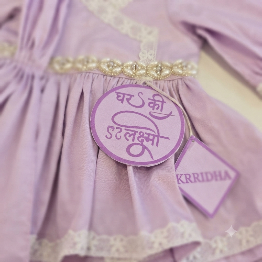 A close-up of a light lavender purple dress with cream lace trim and a decorative beaded waistband. A round purple tag with Hindi text and a smaller tag reading KRRIDHA are attached to the skirt.