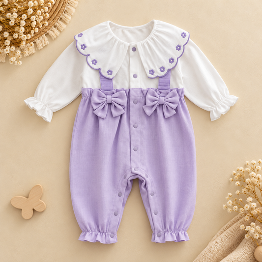 A baby girl's long-sleeve romper in lavender corduroy overalls with a white long-sleeve top underneath, featuring a ruffled lavender collar with embroidered flowers, matching bow details on the suspenders, and snap closures down the front. The romper is displayed in a flat-lay arrangement on a beige background with decorative dried flowers and a wooden butterfly.