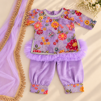A children's traditional outfit laid flat on a light beige surface, featuring a lavender embroidered top with colorful floral motifs, matching flared pants with floral cuffs, and a sheer lavender dupatta with gold fringe.