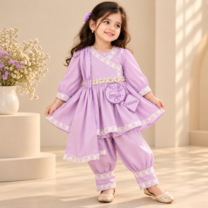 A young girl modeling a light purple two-piece traditional outfit with white lace trim, featuring a long-sleeve tunic and matching pants, in a warm, sunlit indoor setting.