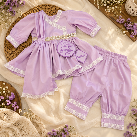 A baby outfit set in light purple/lavender, including a long-sleeve tunic dress with white lace trim and matching bloomers, styled on cream fabric with flowers and a woven tray.