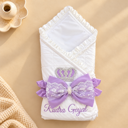 A white quilted baby swaddle wrap with ruffled trim, a lavender bow with white lace, a rhinestone crown embellishment, and the personalized name 'Kudra Goyal' printed in lavender, styled on a beige background.