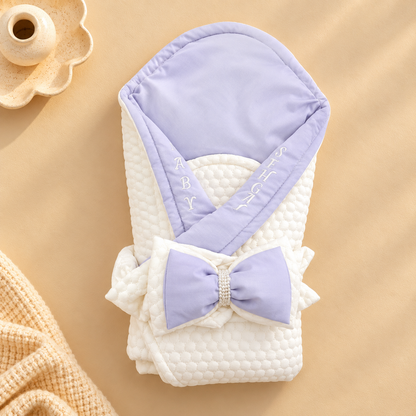 A folded baby blanket or swaddle with a large bow, shown on a warm beige background. The blanket is white with a quilted texture and features a lavender purple hood and matching bow with white embroidered lettering.