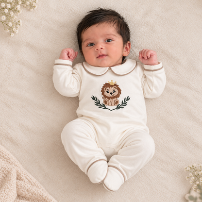 A baby lying on a soft cream blanket wearing an off-white long-sleeve footed romper with a cute embroidered lion graphic on the chest.