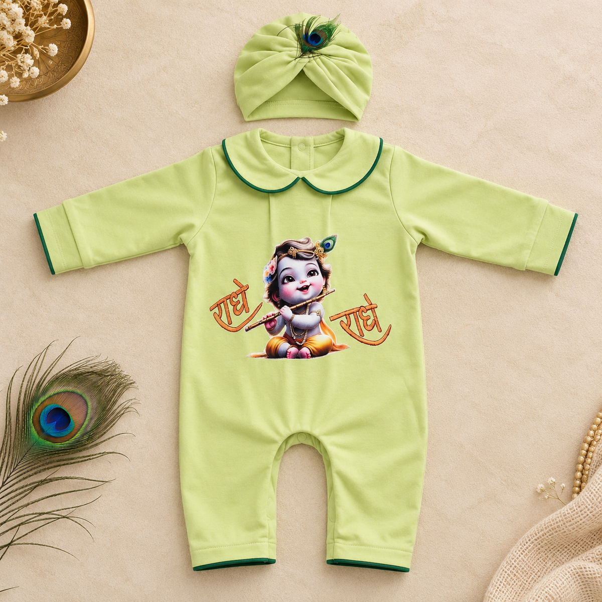 A baby romper and matching turban hat set in light green, styled flat-lay on a beige background with decorative peacock feathers and dried flowers. The romper features a cartoon Krishna graphic with Hindi text 'Radhe Radhe' on the front.