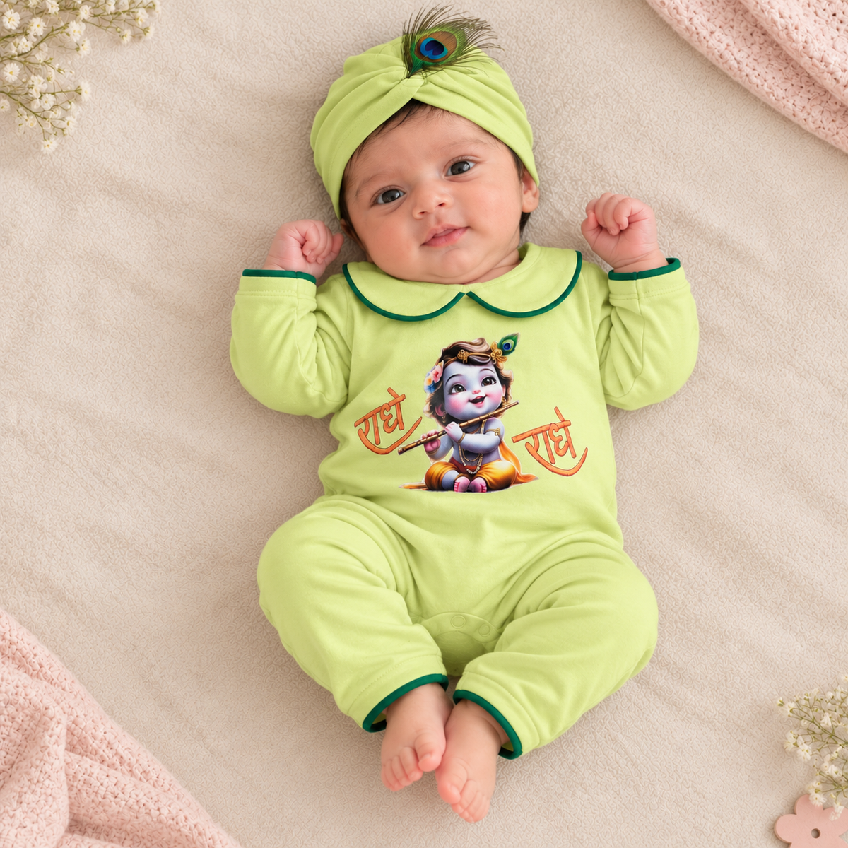 A baby lying on a soft beige blanket wearing a light green long-sleeve romper and matching turban hat with a peacock feather. The romper has a colorful graphic of Lord Krishna and the Hindi text 'राधे राधे' on the front.