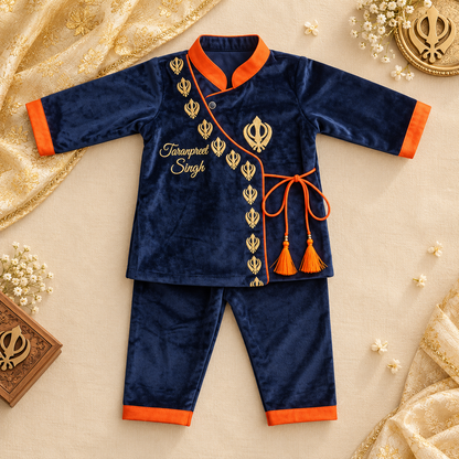 A two-piece baby outfit set in navy blue velvet with bright orange trim, featuring a long-sleeve top with gold embroidery and matching pants. The set is styled on a beige fabric background with decorative gold fabric, small white flowers, and a wooden box.