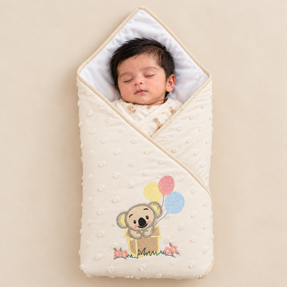 A sleeping newborn baby wrapped in a cream-colored textured muslin swaddle blanket with a cute koala holding balloons design, set against a solid beige background.