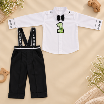 A baby boy's two-piece outfit set featuring a white long-sleeve shirt with a black bow tie, a green sequined number 1 on the chest, and black suspenders with white lettering, laid flat on a beige background with decorative props.