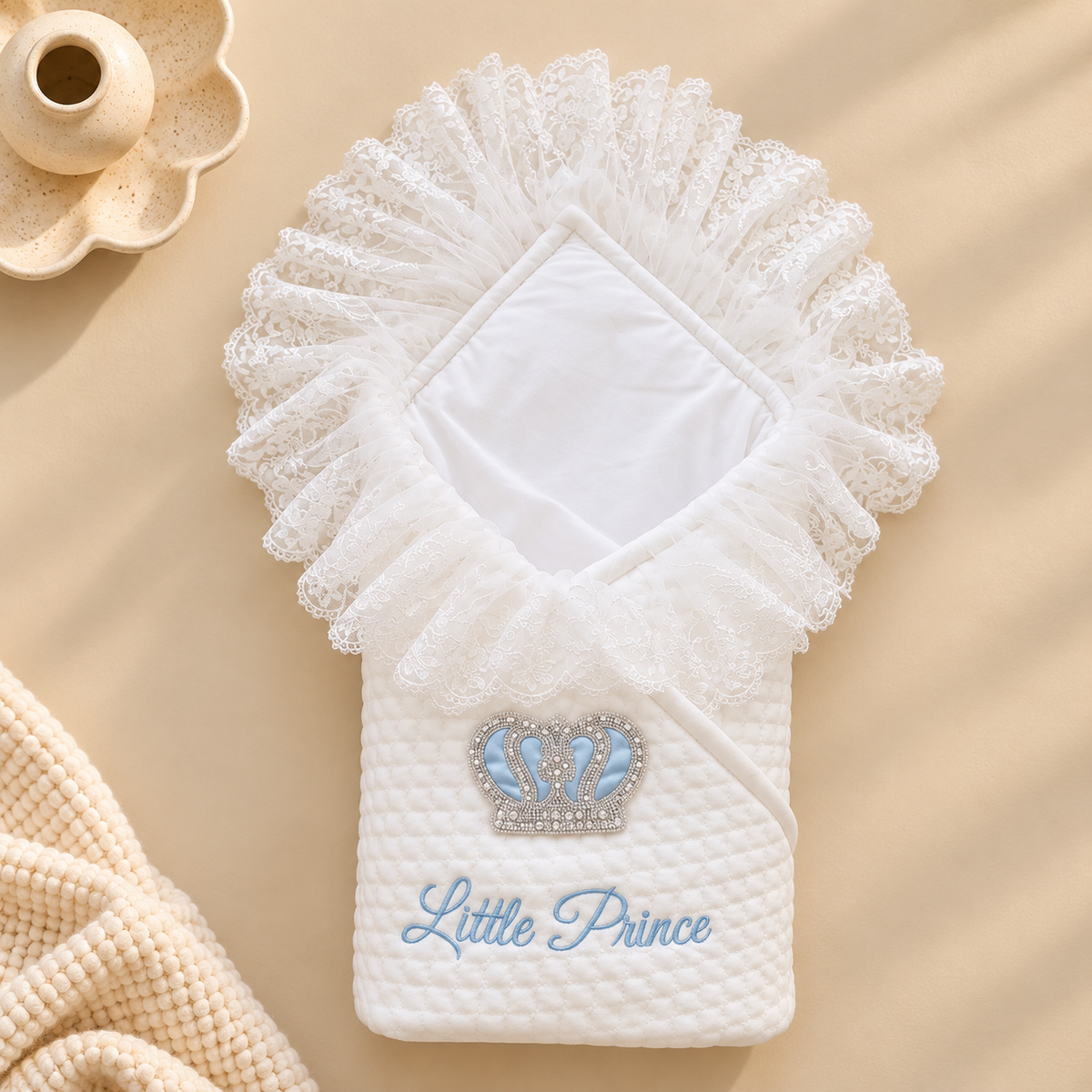 A white quilted baby swaddle blanket with a ruffled lace hood, featuring a rhinestone-embellished blue crown and the words 'Little Prince' embroidered in light blue. The swaddle is laid flat on a beige background with a small decorative ceramic dish in the top left and a cream knit blanket in the bottom left.
