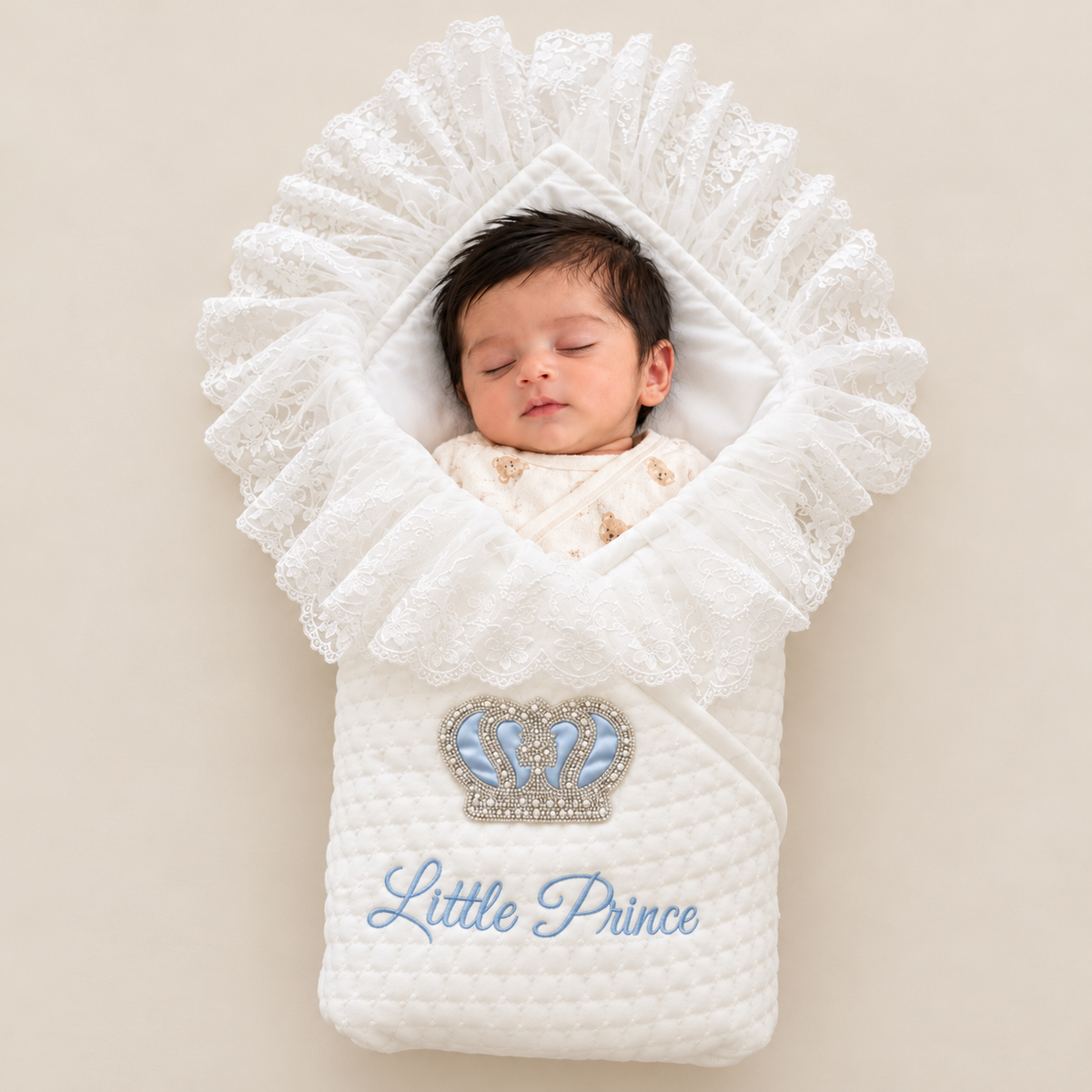 A sleeping newborn baby swaddled in a white quilted wrap blanket with a ruffled lace hood. The blanket features a silver and blue crown graphic and the text 'Little Prince' in light blue embroidery, set against a solid beige background.