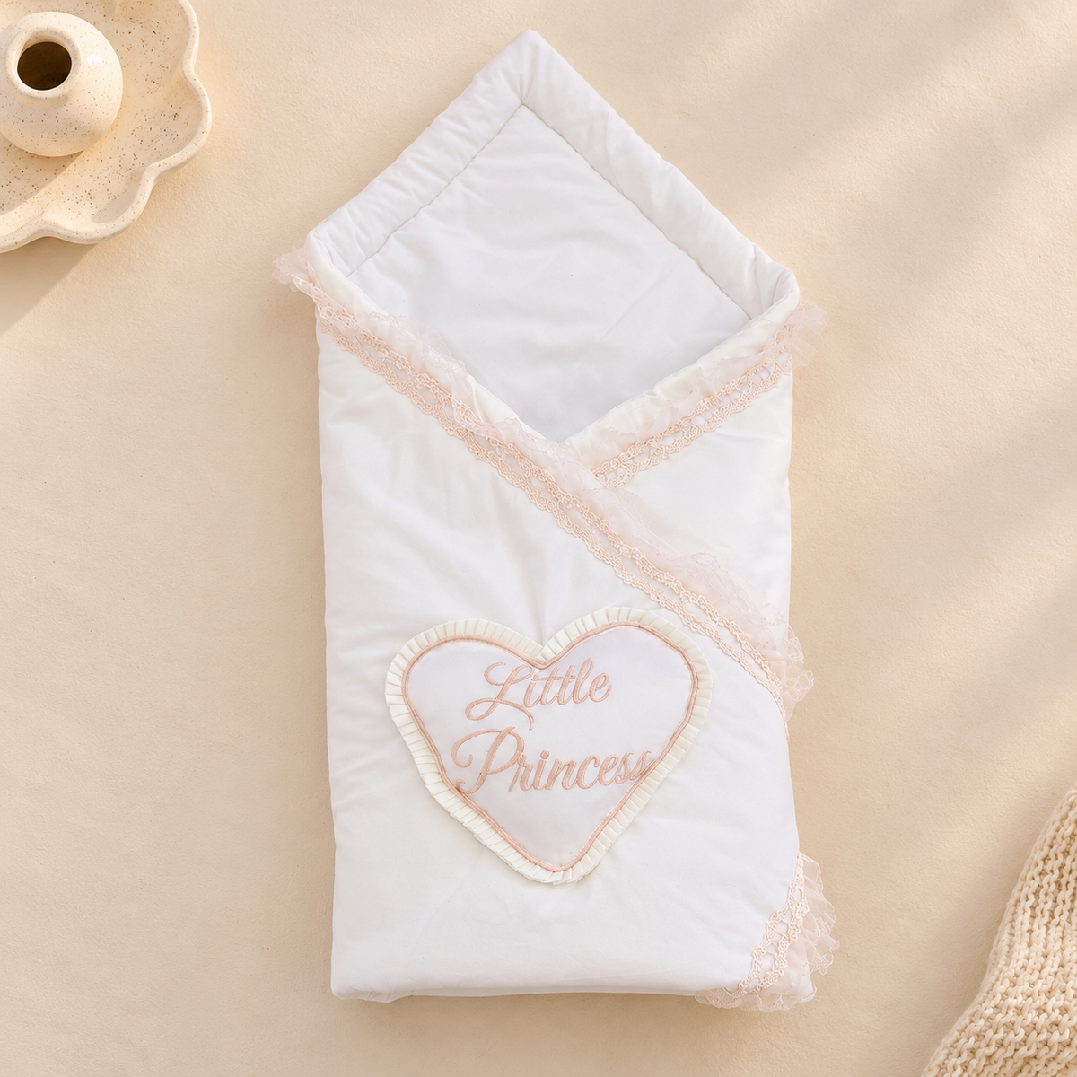 A white baby swaddle blanket with a heart-shaped patch that reads 'Little Princess' in pink, styled on a beige fabric background with decorative props.