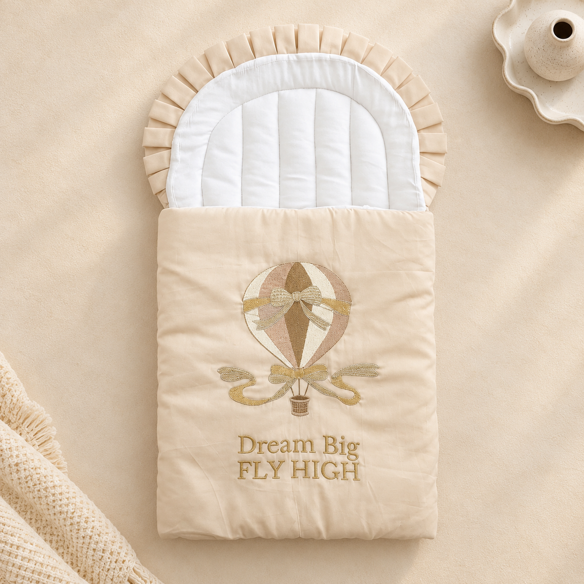 A beige baby sleeping bag or pouch with a white padded interior, featuring an embroidered hot air balloon and the words 'Dream Big FLY HIGH' on the front, styled on a beige fabric background with a textured throw and a small ceramic dish.