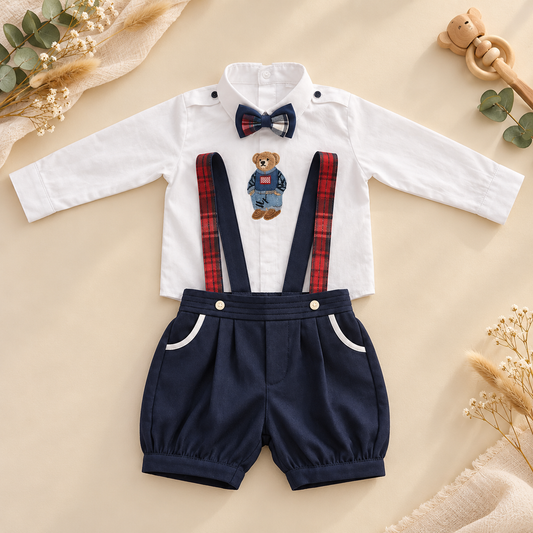 A baby boy's two-piece clothing set featuring a white long-sleeve shirt with a navy bow tie and a teddy bear graphic, paired with navy blue shorts that have red plaid suspenders and white pocket trim, laid flat on a beige background with decorative props.