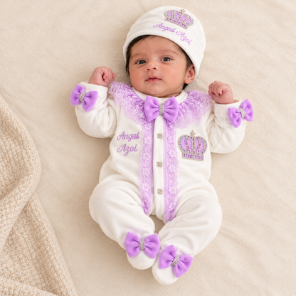 A baby lying on a soft cream blanket wearing a white footed onesie with purple lace trim, purple bows, and a matching white hat with a purple crown and the words 'Angel Ayol'.