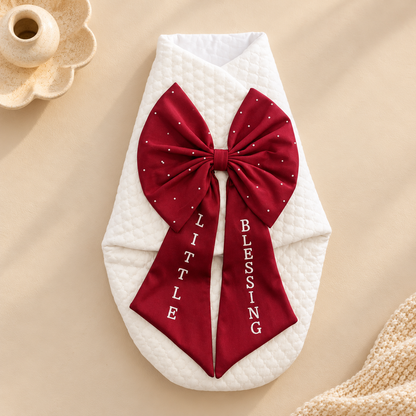 A white quilted baby wrap blanket styled flat-lay with a large deep red satin bow featuring white polka dots and the words 'BLESSED LITTLE BLESSING' printed vertically in white on the tails, set against a warm beige background with decorative props.