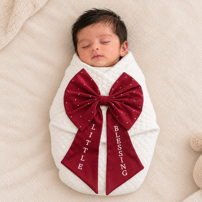 A sleeping newborn baby swaddled in a white quilted blanket with a large burgundy bow featuring white polka dots and the words 'LITTLE BLESSED' in white text.