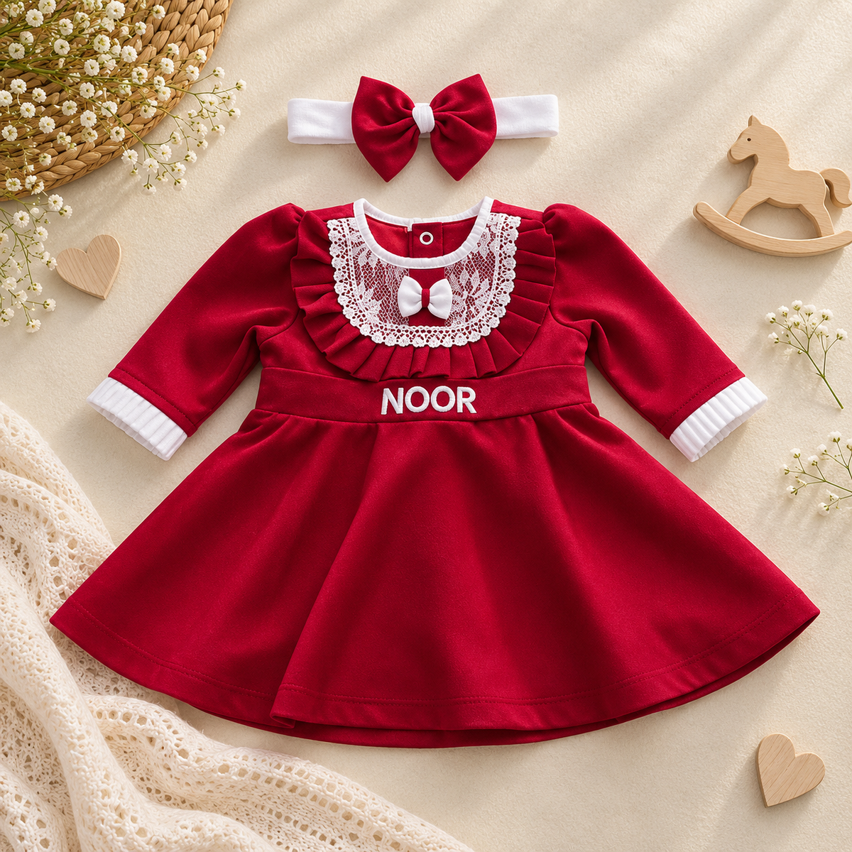 A baby girl's red long-sleeve dress with a white lace collar, white cuffs, and the name 'NOOR' printed on the chest, shown with a matching red and white bow headband. The outfit is styled in a flat lay on a beige fabric background with decorative wooden hearts, a rocking horse, and small white flowers.