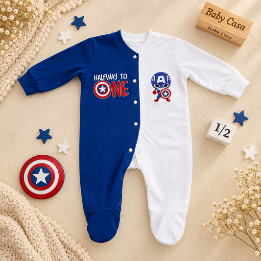 A baby sleeper footed in one-piece, styled in a flat-lay with Captain America themed props including a shield, star stickers, a wooden 'Baby Casa Daily Care' box, and a '1/2' block on a beige background.