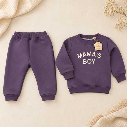 A toddler boy's two-piece sweatshirt and jogger pants set in a dusty purple color, laid flat on a beige background with decorative dried flowers and textured fabric. The sweatshirt features 'MAMA'S BOY' printed in cream across the chest, with a 'baby pride' tag attached.