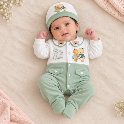 A baby lying on a soft beige blanket wearing a white and sage green one-piece outfit with a matching beanie. The outfit has a white upper section with a green collar, embroidered teddy bear graphics, and the words 'Baby Gupta,' plus sage green pants and matching booties.