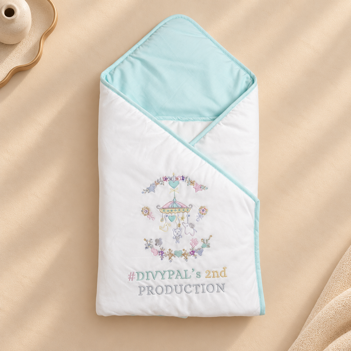 A white baby wrap blanket with a light blue hood and trim, featuring a colorful embroidered carousel design and the text '#DIVYPAL's 2nd PRODUCTION' on a beige fabric background.